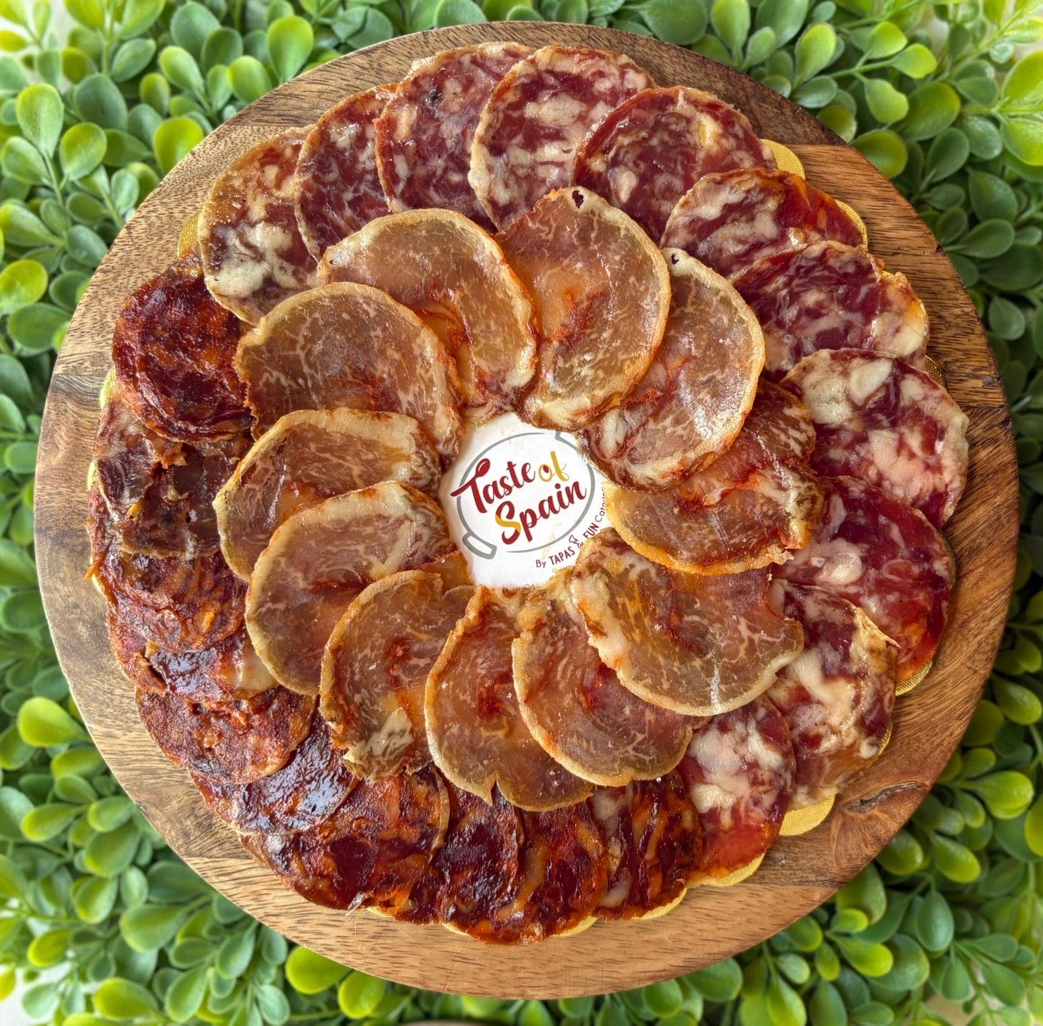 Platter of sliced cured meats on a wooden board with 'Taste of Spain' logo, surrounded by greenery.